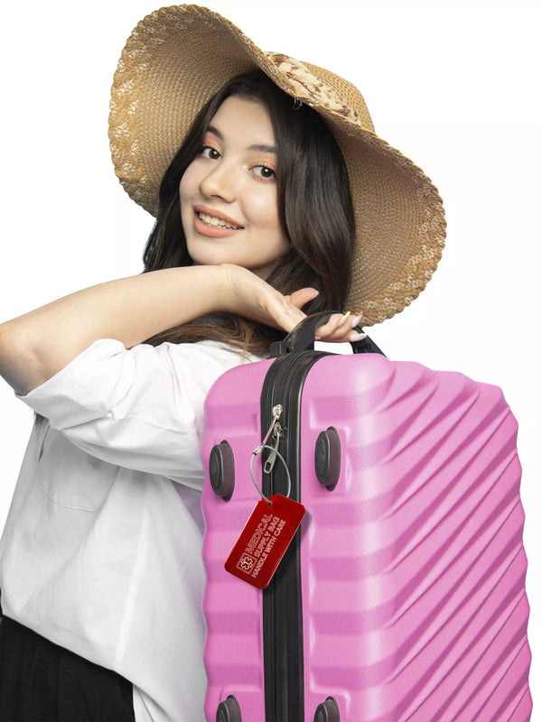 Smiling woman with a hat carrying a pink suitcase with a red 'Medical Supply Bag Handle With Care' luggage tag attached.